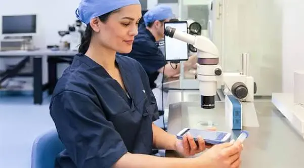 Une technicienne de laboratoire d'AMP, en blouse bleue et charlotte, utilise un lecteur eWitness® pour scanner un tube avant une manipulation sous microscope.